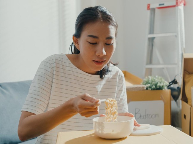 Young woman moving house sit on couch eating instant noodles after unpacking.