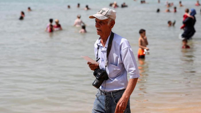 Kisah 50 Tahun Fotografer Mesir dari Hitam-putih ke Digital Egyptian beach photographer Khaled Abdel Rahman, 74, who has been taking pictures of couples and families frequenting the shores of the Suez Canal for over 50 years, even after the rise in popularity of mobile photography, shows people their photos during a hot day at a public beach at the entrance of the Suez Canal, Egypt, September 13, 2024. REUTERS/Mohamed Abd El Ghany