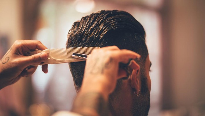 Close-up of barbers tattooed hands holding comb and scissors and giving man trendy hairstyle