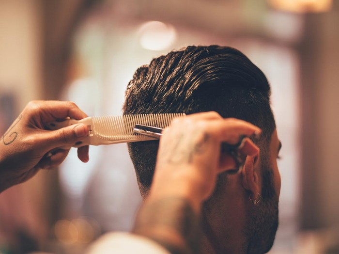 Close-up of barbers tattooed hands holding comb and scissors and giving man trendy hairstyle