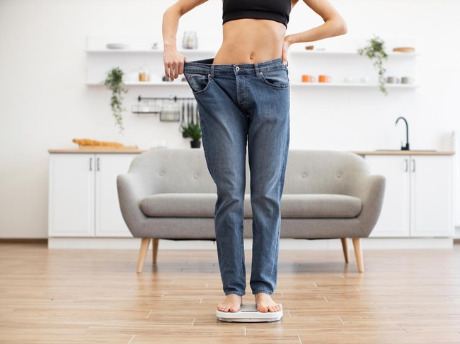 Woman standing on scale with loose jeans, demonstrating significant weight loss success, in modern living room. Healthy lifestyle and fitness achievement concept.