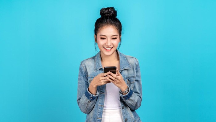 Portrait of Happy asian woman feeling happiness and standing typing smartphone on blue background. Cute asia girl smiling wearing casual jeans shirt and connect internet shopping online and surfing.