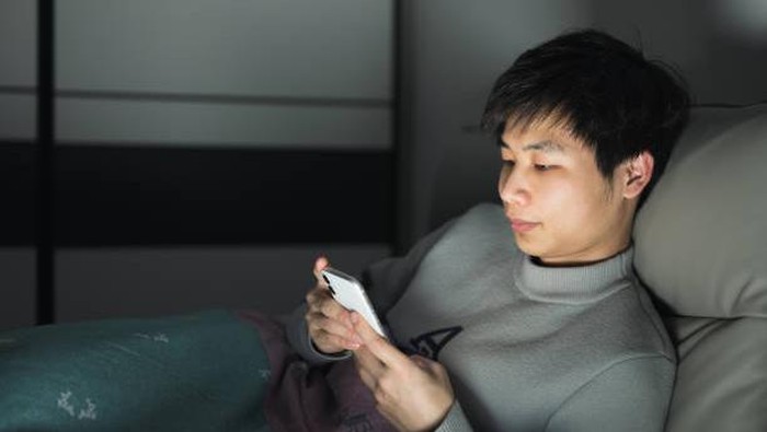 At night, a young Asian man sits on his bed and plays with his mobile phone, the screen light illuminating his face.