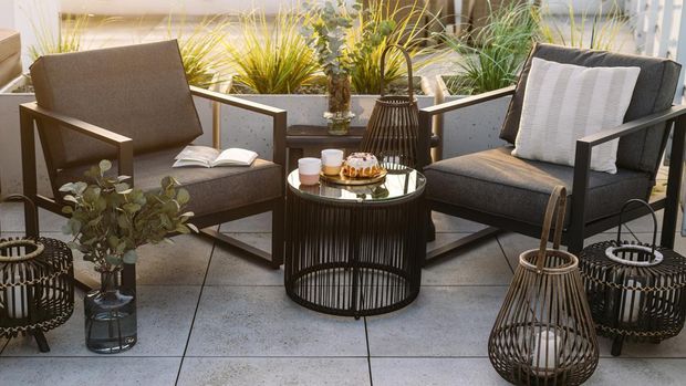 Two modern patio chairs close to evening lanterns on balcony. Armchairs near glass top table on roof terrace with green potted plants. Residential building with cityscape view on background