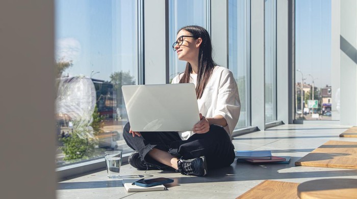 Wanita sedang duduk di pinggir jendela sambil memegang laptop dan duduk dengan nyaman