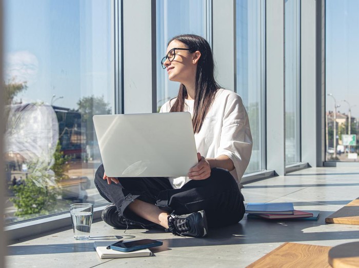 Wanita sedang duduk di pinggir jendela sambil memegang laptop dan duduk dengan nyaman