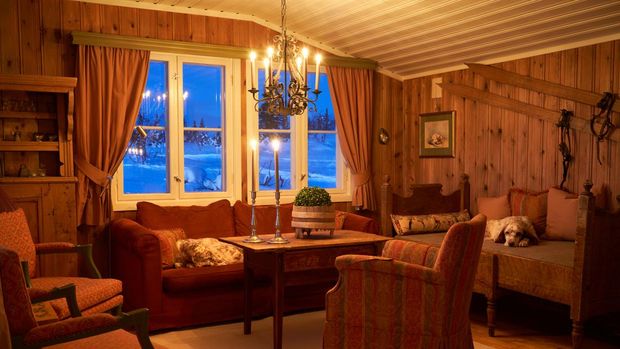Living room interior at dusk lit by candlelight with two English setters sleeping, mountain landscape with deep snow visible through the window. Norway