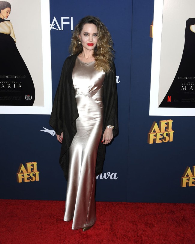 Jolie yang tampil ala Old Hollywood Glam, terlihat elegan dalam balutan gaun sutra berwarna sampanye yang memiliki kerah tinggi dan detail dart atau lipatan kupnat pada bagian dada. Dia melengkapi penampilannya dengan selendang hitam, anting-anting mutiara dan gelang yang serasi dengan busananya. Foto: Getty Images