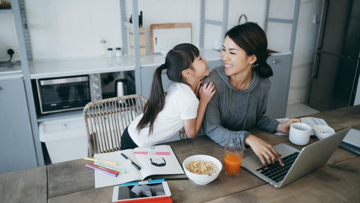 Young Asian mother working from home on a laptop while little daughter is studying from home with a digital tablet. They are enjoying the time together