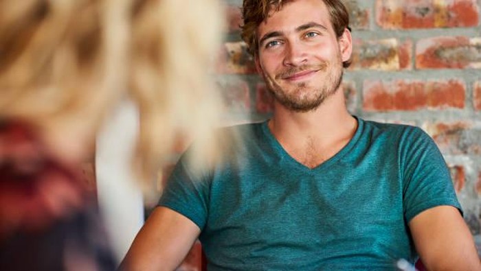 Loving young man looking at woman. Handsome male is sitting against brick wall. He is wearing t-shirt at home.