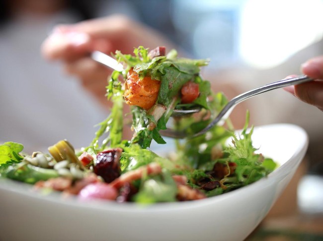 Ilustrasi makan salad untuk diet A cropped image of an Asian woman mixing ingredients in her healthy fresh vegan salad. The close-up showcases a multi-colored salad bowl filled with assorted fresh leaf vegetables, including rocket, tomato, zucchini, olives and pumpkin seeds.