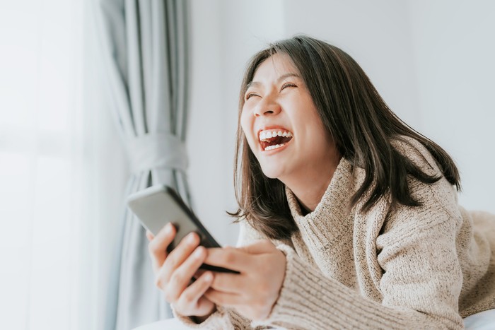 Happy Asian woman laughing while lying on the bed using smartphone mobile at home
