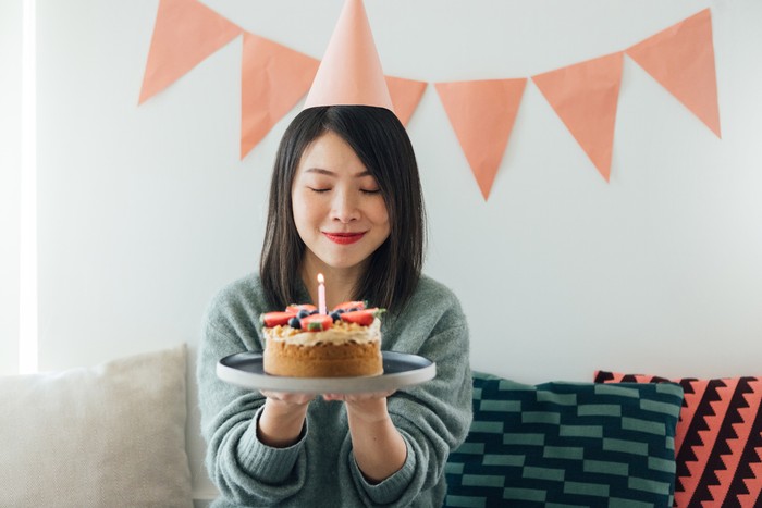Portrait of beautiful young Asian woman with party hat waking a wish while holding a strawberry birthday cake, having a birthday party at home with pink coloured bunting hanging on a white wall.