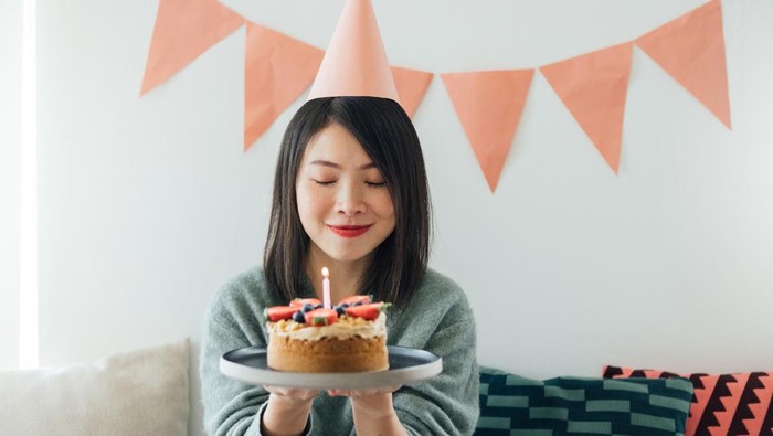 Portrait of beautiful young Asian woman with party hat waking a wish while holding a strawberry birthday cake, having a birthday party at home with pink coloured bunting hanging on a white wall.