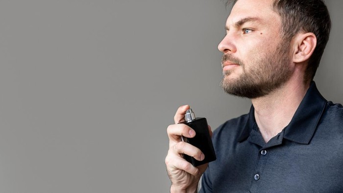 Applying perfume. Man spraying cologne on his body in front of gray plain background with copy space.
