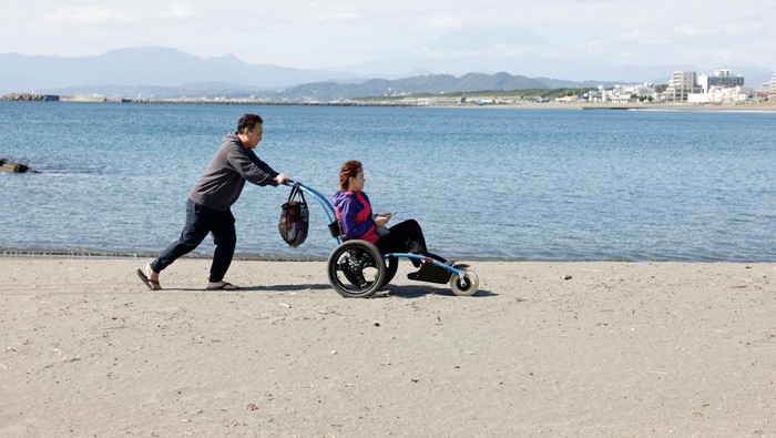 Ahli Manikur Jepang Olah Limbah Plastik Jadi Kuku Palsu Cantik Japanese manicurist Naomi Arimoto's husband pushes her in a wheelchair while she collects plastic waste from the sand at a beach in Chigasaki, Kanagawa Prefecture, Japan October 21, 2024. REUTERS/Manami Yamada