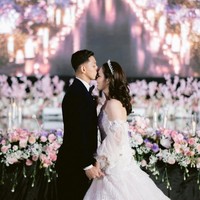 Berbeda dengan akad nikah yang mengusung tema tradisional, resepsi Rian dan Ribka bergaya internasional. Rian tampil gagah mengenakan jas navy berbahan velvet dilengkapi dasi kupu-kupu, sementara Ribka tampil bak princess dengan gaun pengantin putih. Foto: Instagram/@ribkasugiarto