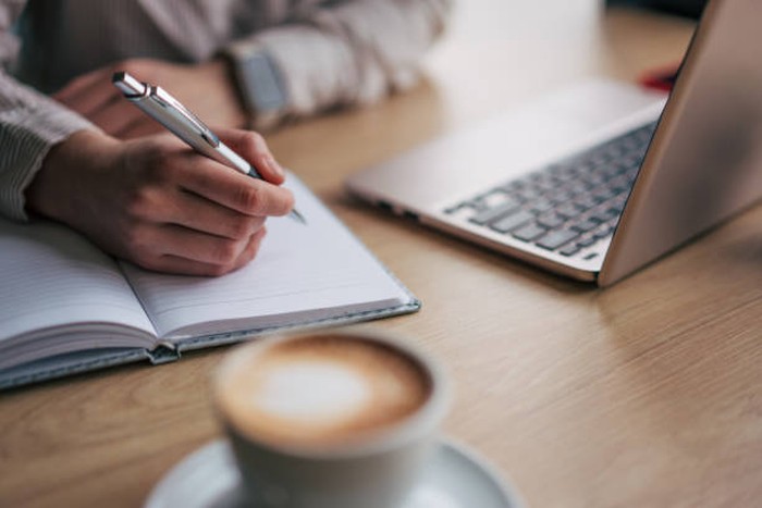Close up photo of female hand writting down something. There is laptop behind the notebook.