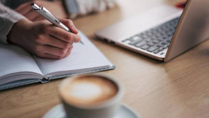 Close up photo of female hand writting down something. There is laptop behind the notebook.