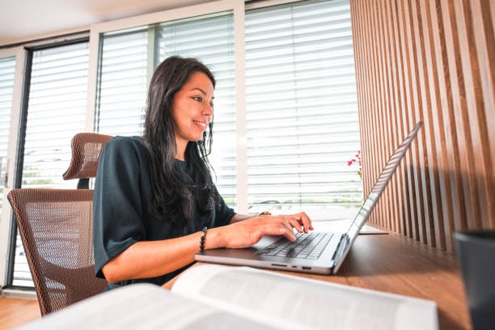 Hispanic woman sitting at her desk, using a laptop and typing. She is wearing casual clothes.