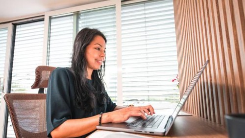 Hispanic woman sitting at her desk, using a laptop and typing. She is wearing casual clothes.