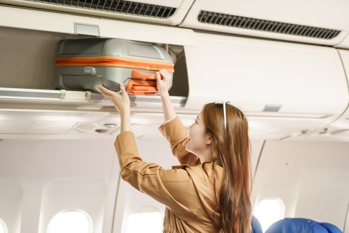 An Asian woman travels abroad by plane to study. She stands on the plane to collect her luggage, smiling and happy with the journey, excited about her new educational adventure.