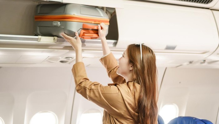 An Asian woman travels abroad by plane to study. She stands on the plane to collect her luggage, smiling and happy with the journey, excited about her new educational adventure.