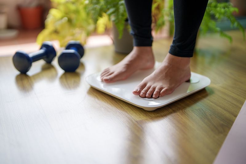 A close-up view of a young woman standing on a weighing scale, focusing on her feet and the display as she checks her weight. The scene highlights the importance of health and self-awareness in maintaining a balanced lifestyle.