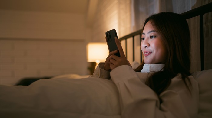 Portrait of Asian teen girl lying in bed at night and using smartphone. Young woman lying in bed using smartphone for social media and watching series at late night. Insomnia, sleep disorder concept.