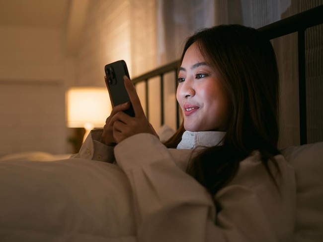 Portrait of Asian teen girl lying in bed at night and using smartphone. Young woman lying in bed using smartphone for social media and watching series at late night. Insomnia, sleep disorder concept.