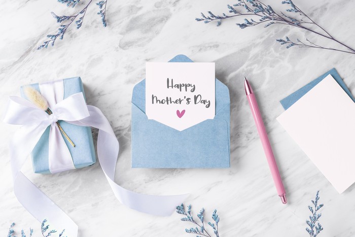 Mothers day card with blue gift box and envelope on marble table with flower and pen, top view