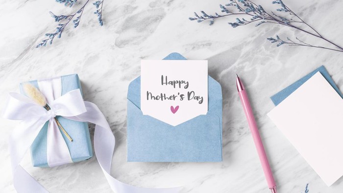 Mothers day card with blue gift box and envelope on marble table with flower and pen, top view