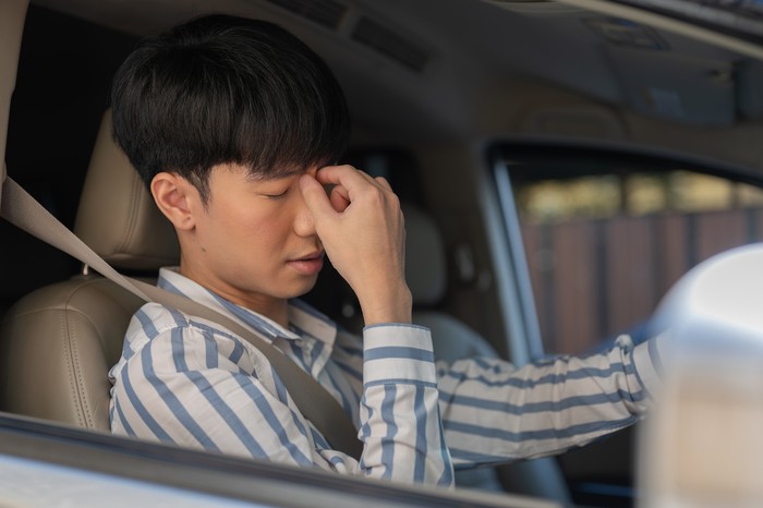 A tired young-adult Asian man is suffering from headaches or eye strain while driving a car for long hours. People and vehicle concepts
