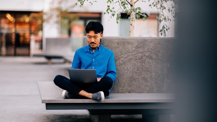 A young man in a blue shirt works on his laptop while sitting cross-legged outdoors, embodying focus and relaxation in a public urban area.