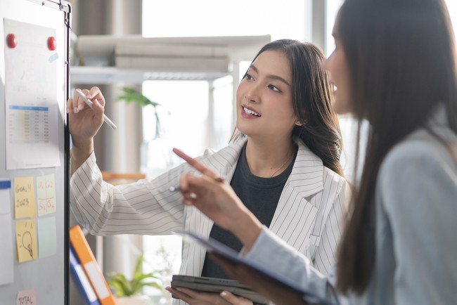 two Businesswoman brainstorming ideas on whiteboard with colleague. Female business partners having brainstorming session about financial budget in startup meeting room.