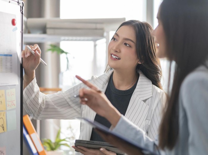 two Businesswoman brainstorming ideas on whiteboard with colleague. Female business partners having brainstorming session about financial budget in startup meeting room.