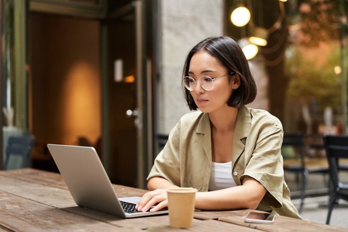 Young woman working in a cafe, using laptop and drinking coffee. Asian girl student with computer studying remotely, sitting on bench near shop.