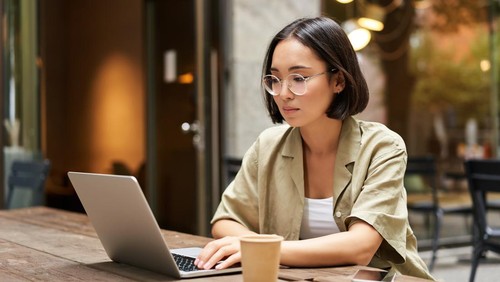 Young woman working in a cafe, using laptop and drinking coffee. Asian girl student with computer studying remotely, sitting on bench near shop.
