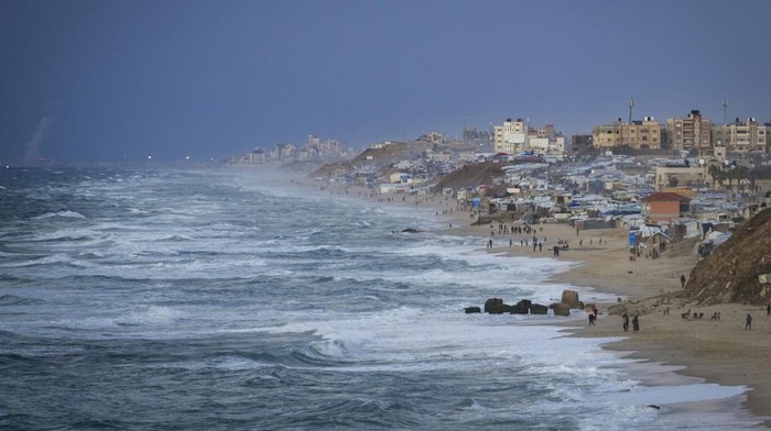 People walk along the beach next to a tent refugee camp for displaced Palestinians in Deir al-Balah, central Gaza Strip, Monday, Dec .30, 2024. (AP Photo/Abdel Kareem Hana)