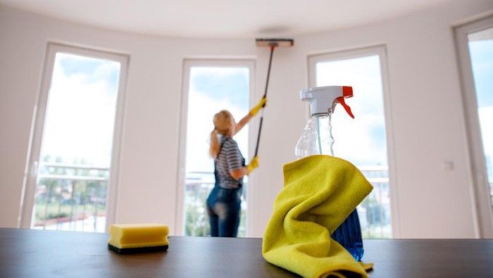 Yellow cleaning rag and sponge with spraying bottle on desk with blurred housewife cleaning the ceiling in the background. House cleaning concept. Copy space