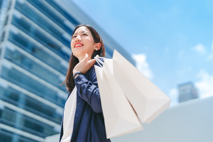 People city lifestyles with consumerism. Adult asian business woman happy smiling face standing at outdoor with shopping bags. City building background with blue sky.