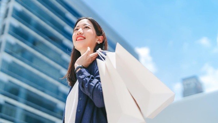People city lifestyles with consumerism. Adult asian business woman happy smiling face standing at outdoor with shopping bags. City building background with blue sky.