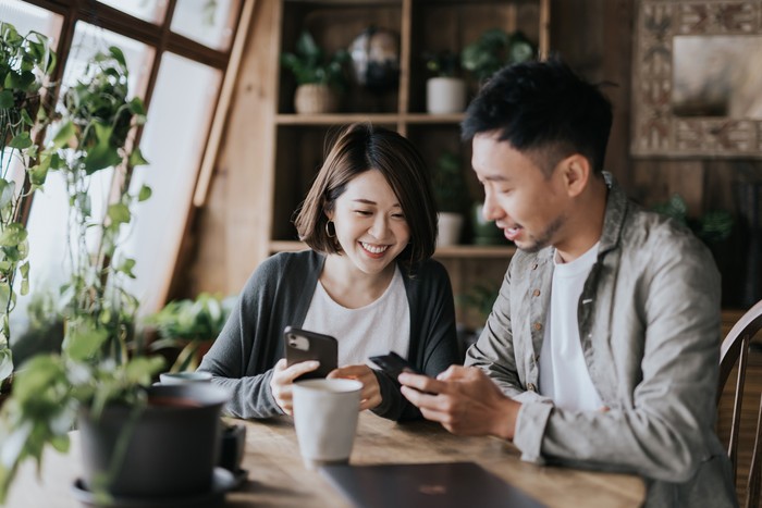 Pasangan Happy young Asian couple having a coffee date in cafe. Drinking coffee and chatting while using smartphone together. Enjoying a relaxing moment