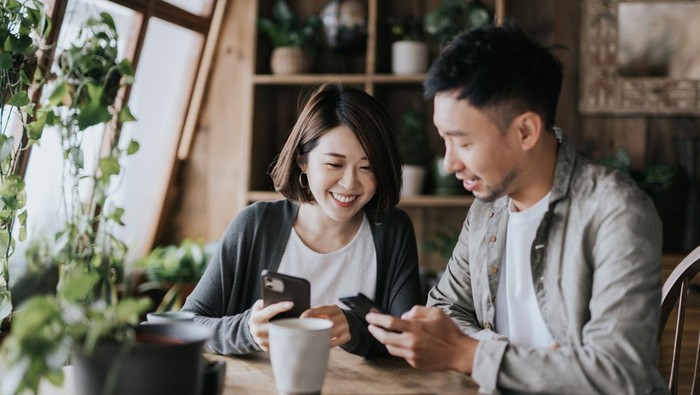 Happy young Asian couple having a coffee date in cafe. Drinking coffee and chatting while using smartphone together. Enjoying a relaxing moment