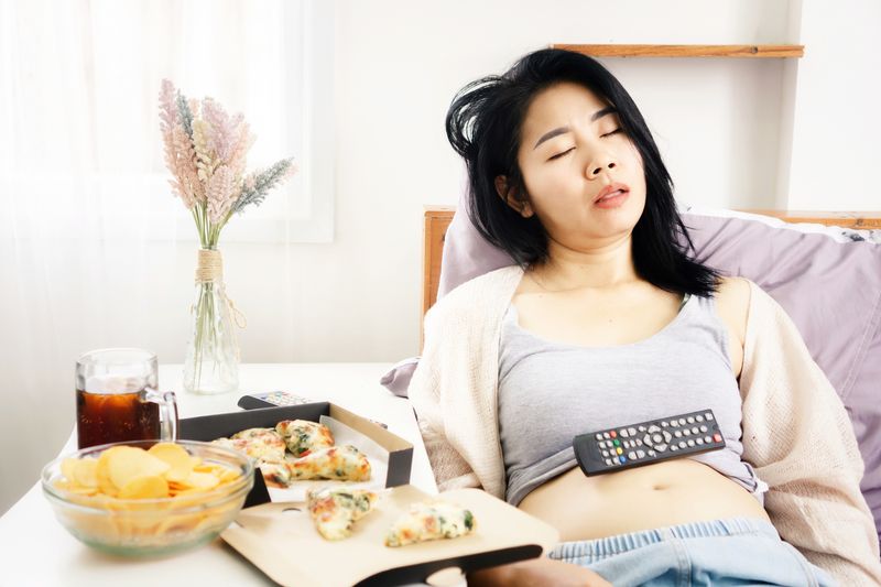 ngantuk Asian woman sleeping in bed after eating pizza, potato chips and soda with Tv remote on her fat belly overeating junk food concept