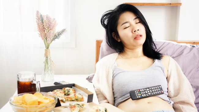 Asian woman sleeping in bed after eating pizza, potato chips and soda with Tv remote on her fat belly  overeating junk food concept