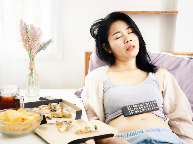 Asian woman sleeping in bed after eating pizza, potato chips and soda with Tv remote on her fat belly  overeating junk food concept