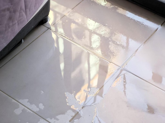 trembling water puddle after pipe burst covers white tile near furniture in room reflecting window and blue sky close view