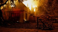 Api membumbung dari sebuah rumah saat sebuah sepeda roda tiga terparkir di halaman, sementara angin kencang yang memicu kebakaran hutan dahsyat di wilayah Los Angeles memaksa warga mengungsi, di Kebakaran Eaton di Altadena, California, AS, 8 Januari 2025. 