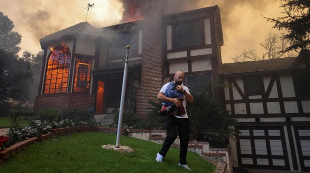 Seorang yang membawa bendera berjalan menjauh dari rumah yang terbakar saat angin kencang yang memicu kebakaran hutan yang dahsyat di wilayah Los Angeles memaksa orang-orang untuk mengungsi, di Kebakaran Eaton di Altadena, California, AS, 8 Januari 2025. (REUTERS/David Swanson)